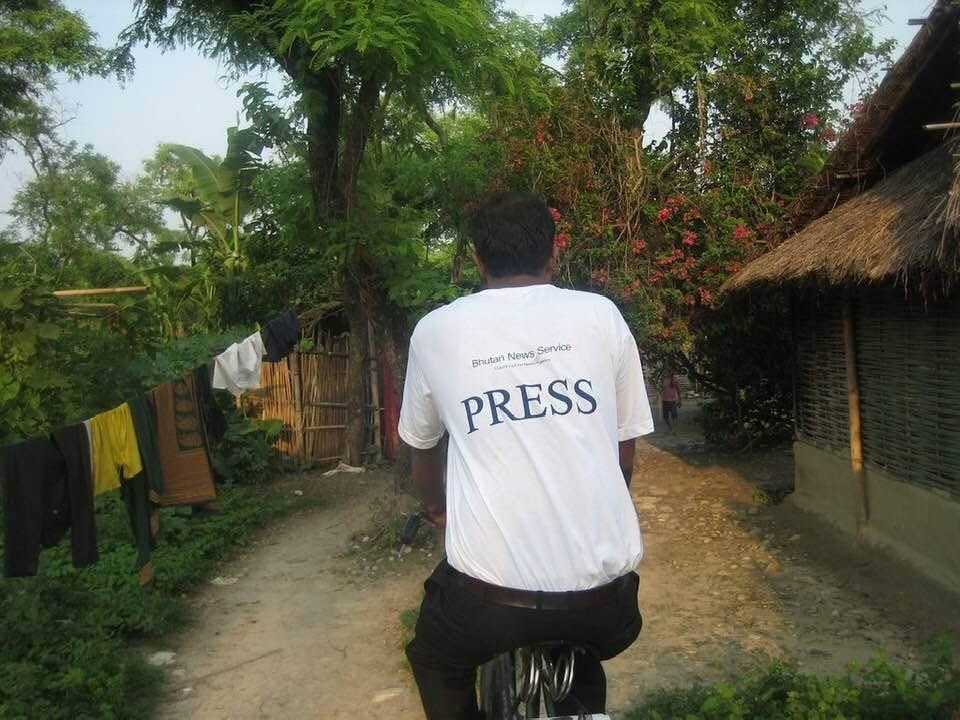 TP Mishra rides a bicycle through a refugee camp wearing a Bhutan News Service PRESS shirt